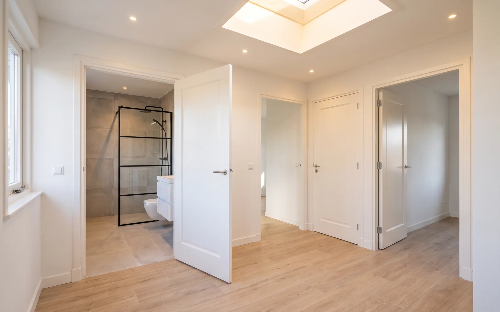 Een net verbouwde overloop met een nieuwe vloer, een strak plafond en door de open deur een nieuwe badkamer met inloopdouche